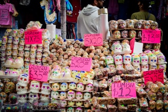 A vibrant street market stall displaying an array of colorful skull-shaped sugar candies, commonly known as sugar skulls, associated with the Day of the Dead tradition in Mexico. Bright pink signage indicates the pricing for the various items. The background includes hanging clothing items, and a few people are seen browsing or standing nearby.