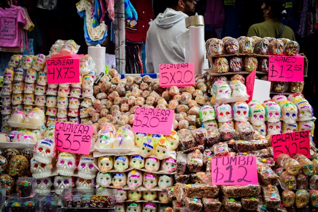 A vibrant street market stall displaying an array of colorful skull-shaped sugar candies, commonly known as sugar skulls, associated with the Day of the Dead tradition in Mexico. Bright pink signage indicates the pricing for the various items. The background includes hanging clothing items, and a few people are seen browsing or standing nearby.