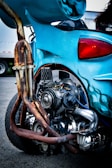 A close-up of a custom motorcycle showcasing its intricately designed exhaust system and engine. The vehicle features a vibrant blue color with flame decals, and parts show signs of rust and wear. The focus is on the mechanical details and craftsmanship of the bike.