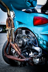 A close-up of a custom motorcycle showcasing its intricately designed exhaust system and engine. The vehicle features a vibrant blue color with flame decals, and parts show signs of rust and wear. The focus is on the mechanical details and craftsmanship of the bike.