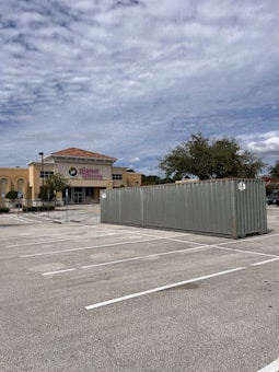 A large shipping container is placed in an empty parking lot. In the background, there is a building with a sign reading 'Planet Fitness' and a few trees. The sky is cloudy with a mix of gray and blue tones.