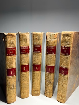 Five antique hardcover books with ornate golden spines are lined up side by side. The spines are adorned with red labels displaying the text 'Théâtre de Racine' and numbered from 1 to 5. The books show signs of wear, with some fraying and fading, indicating age and use.