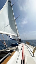A sailboat navigating through calm waters with its white sails unfurled against a partly cloudy sky. The deck consists of metal railings and wooden trim, with ropes neatly organized. The scene has a sense of tranquility and freedom.