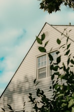 A triangular section of a house with white siding is visible, partially framed by out-of-focus green leaves in the foreground. A window is centered in the middle of the wall segment. The sky is partially cloudy, casting soft shadows on the siding.