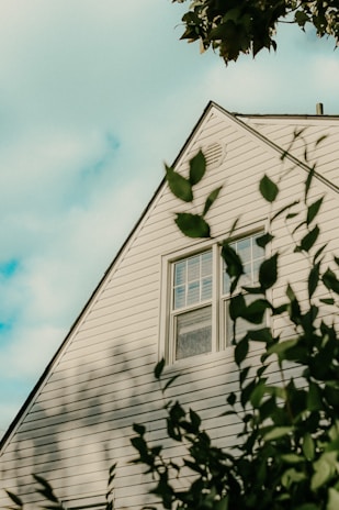 A triangular section of a house with white siding is visible, partially framed by out-of-focus green leaves in the foreground. A window is centered in the middle of the wall segment. The sky is partially cloudy, casting soft shadows on the siding.