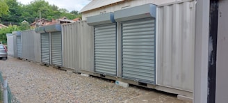 A row of metal shipping containers is converted into storage units with closed roll-up doors. The containers are placed on a gravel path with a small strip of pavement. In the background, there are trees and a partially visible car.