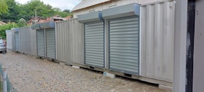 A row of metal shipping containers is converted into storage units with closed roll-up doors. The containers are placed on a gravel path with a small strip of pavement. In the background, there are trees and a partially visible car.