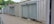 A row of metal shipping containers is converted into storage units with closed roll-up doors. The containers are placed on a gravel path with a small strip of pavement. In the background, there are trees and a partially visible car.