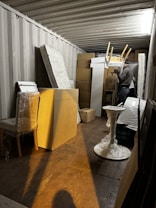 A storage container filled with various items including wrapped furniture and stacked boxes. A person is seen organizing the contents, holding a chair above other items.