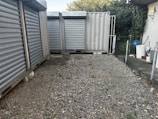 A gravel-covered area enclosed by metal storage containers with closed, corrugated steel doors. There is a chain-link fence on the right, leading to a small area with vegetation. On the left, several large metal containers are aligned. In the background, partial greenery with a hint of the sky is visible.