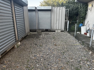 A gravel-covered area enclosed by metal storage containers with closed, corrugated steel doors. There is a chain-link fence on the right, leading to a small area with vegetation. On the left, several large metal containers are aligned. In the background, partial greenery with a hint of the sky is visible.