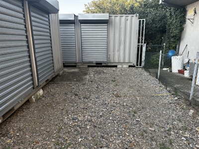 A gravel-covered area enclosed by metal storage containers with closed, corrugated steel doors. There is a chain-link fence on the right, leading to a small area with vegetation. On the left, several large metal containers are aligned. In the background, partial greenery with a hint of the sky is visible.