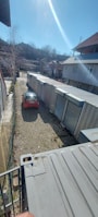 A long row of industrial storage units with metal doors lines one side of a gravel pathway. A red car is parked on the left, partially illuminated by sunlight. Buildings with sloped roofs are in the background, and leafless trees suggest a winter or early spring setting.