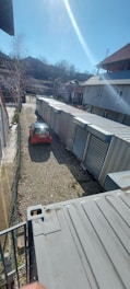 A long row of industrial storage units with metal doors lines one side of a gravel pathway. A red car is parked on the left, partially illuminated by sunlight. Buildings with sloped roofs are in the background, and leafless trees suggest a winter or early spring setting.