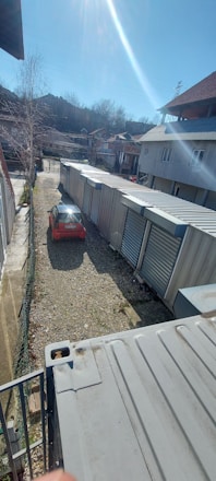 A long row of industrial storage units with metal doors lines one side of a gravel pathway. A red car is parked on the left, partially illuminated by sunlight. Buildings with sloped roofs are in the background, and leafless trees suggest a winter or early spring setting.