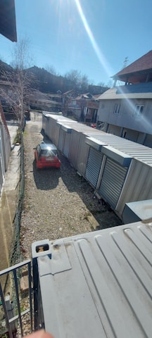 A long row of industrial storage units with metal doors lines one side of a gravel pathway. A red car is parked on the left, partially illuminated by sunlight. Buildings with sloped roofs are in the background, and leafless trees suggest a winter or early spring setting.