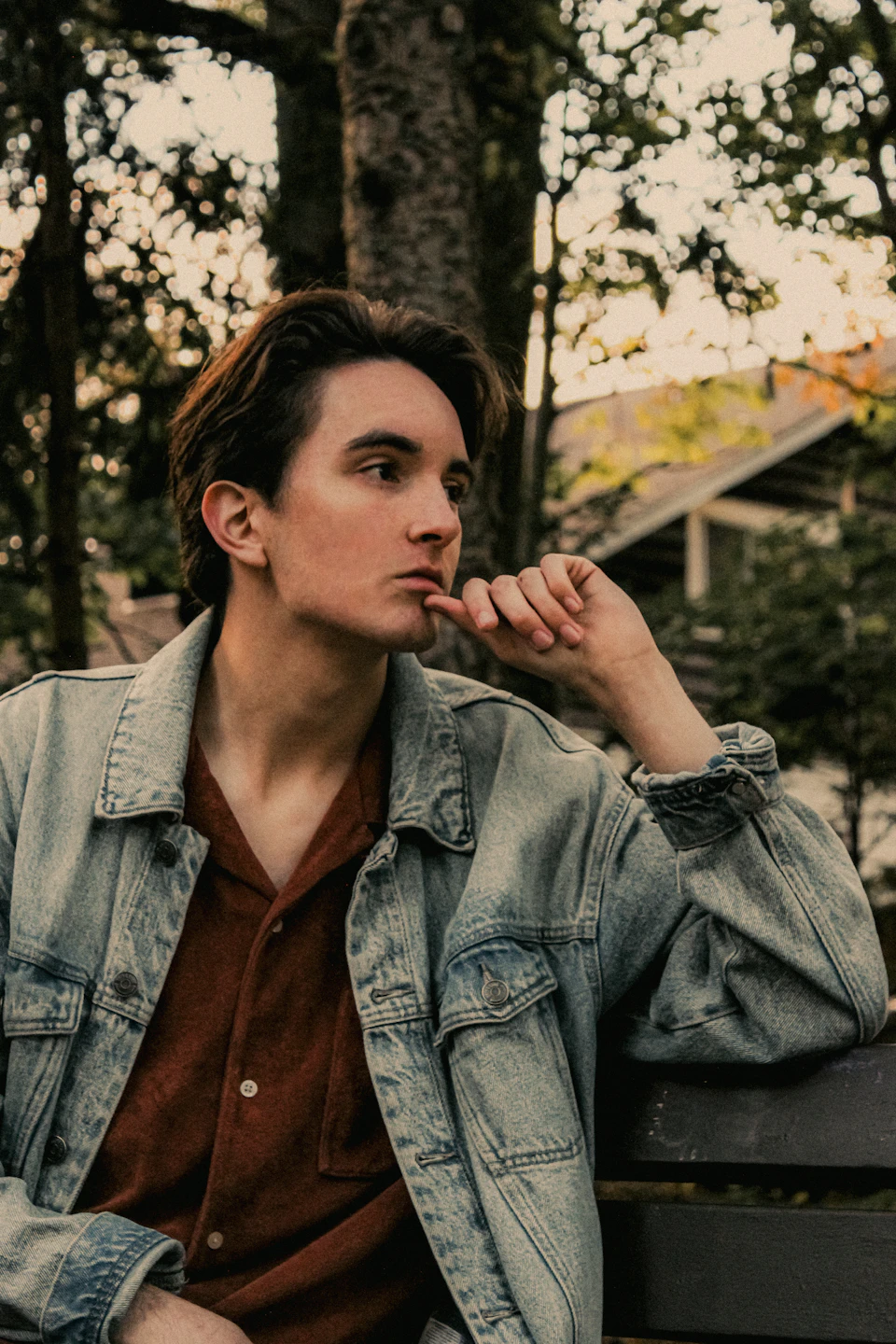 In a serene park setting, a young man is captured in a moment of contemplation. He's dressed casually for the day, wearing a denim jacket over an orange shirt and sporting brown hair that falls into his eyes. His left hand rests on his chin, suggesting deep thought or introspection. The bench he's sitting on is positioned against a backdrop of trees, providing a sense of tranquility to the scene.