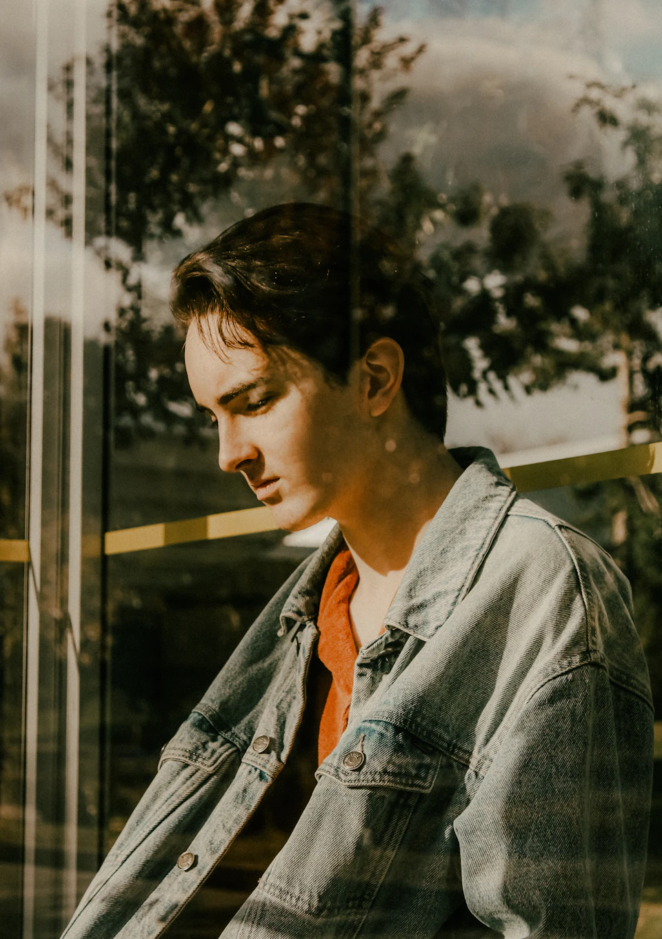 Young man with short brown hair stands against a window reflecting the outdoors landscape