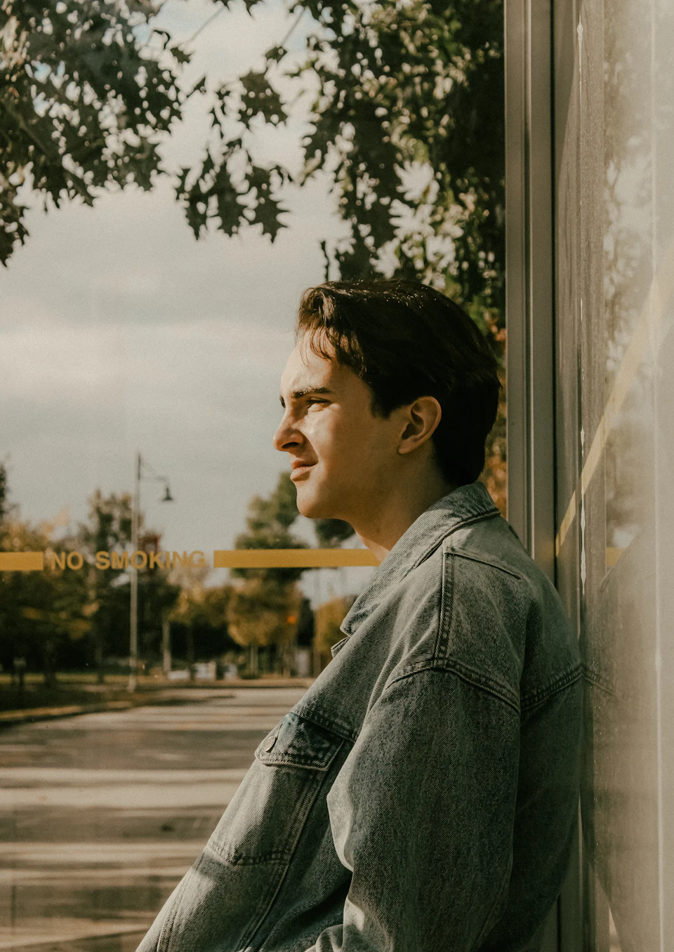 Young man leaning against window on building, wearing denim jacket and contemplating