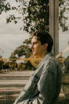 A person is leaning against a reflective glass wall, looking contemplatively into the distance. The scene is outdoors with trees and a sidewalk visible in the background. The person is wearing a denim jacket, and the lighting suggests a warm, sunny day.