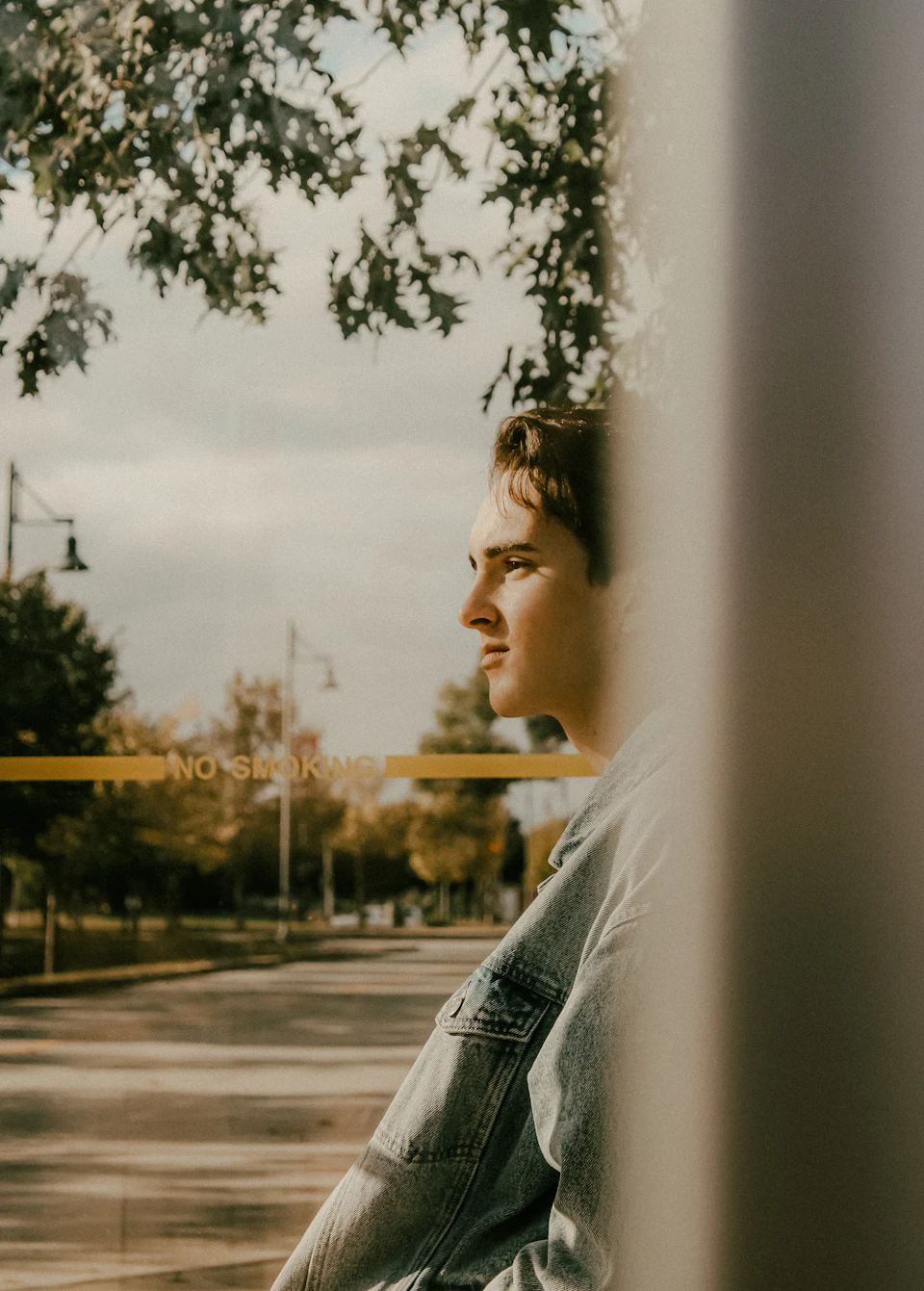 Young man stands on sidewalk with contemplative expression, denim jacket and city trees behind him