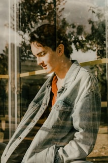 A person wearing a denim jacket and orange shirt sits against a reflective glass surface, with trees and sky faintly visible in the reflection. The lighting creates a serene, contemplative mood.