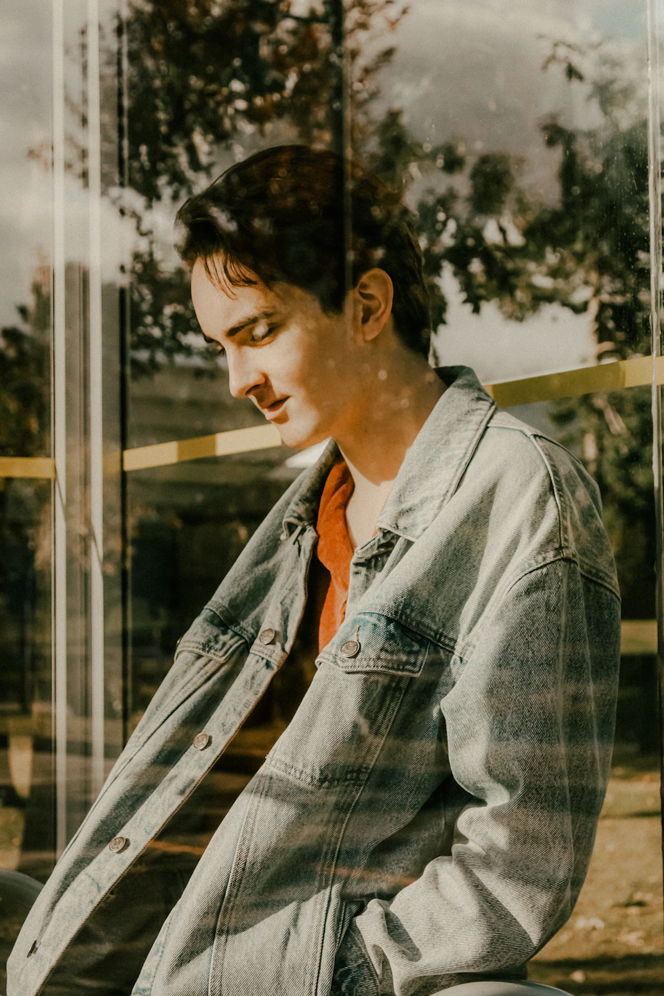 Young man wears denim jacket and shirt, arms crossed, in reflective window scene outdoors