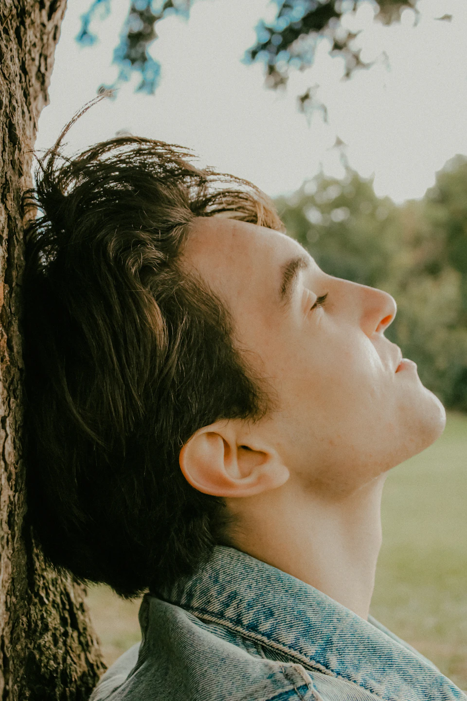 Young man leans against tree trunk, head tilted back, eyes closed in relaxation outdoors