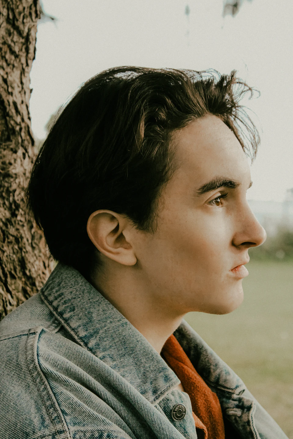 The image shows a young man with short dark hair wearing a denim jacket and an orange shirt, leaning against a tree trunk on what appears to be a grassy field or park setting. The man's gaze is directed off to the side, suggesting he may be lost in thought or simply enjoying his surroundings.
