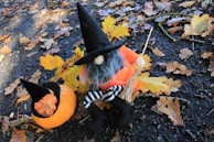 A whimsical doll dressed as a witch sits among fallen autumn leaves on a dark, wet pavement. It wears a tall black hat, has a gray beard, and holds a small broomstick. Beside it, a small pumpkin adorned with a similar witch hat rests among the leaves.