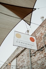 A sign for 'Component Coffee Lab' hangs on a metal pole against the backdrop of a brick wall. The sign features three circular, stylized designs in beige, light yellow, and orange. Above the sign, several stretched fabric shades and hanging light bulbs are visible, hinting at an outdoor seating area.