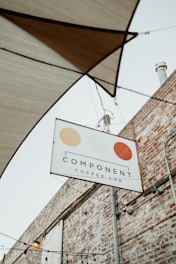 A sign for 'Component Coffee Lab' hangs on a metal pole against the backdrop of a brick wall. The sign features three circular, stylized designs in beige, light yellow, and orange. Above the sign, several stretched fabric shades and hanging light bulbs are visible, hinting at an outdoor seating area.