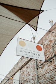 A sign for 'Component Coffee Lab' hangs on a metal pole against the backdrop of a brick wall. The sign features three circular, stylized designs in beige, light yellow, and orange. Above the sign, several stretched fabric shades and hanging light bulbs are visible, hinting at an outdoor seating area.
