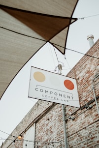 A sign for 'Component Coffee Lab' hangs on a metal pole against the backdrop of a brick wall. The sign features three circular, stylized designs in beige, light yellow, and orange. Above the sign, several stretched fabric shades and hanging light bulbs are visible, hinting at an outdoor seating area.