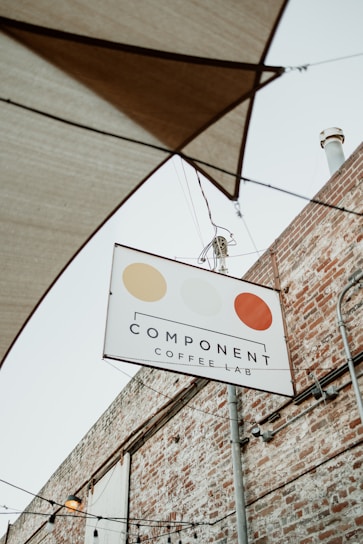 A sign for 'Component Coffee Lab' hangs on a metal pole against the backdrop of a brick wall. The sign features three circular, stylized designs in beige, light yellow, and orange. Above the sign, several stretched fabric shades and hanging light bulbs are visible, hinting at an outdoor seating area.