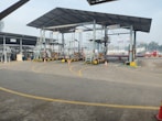 An industrial setting with multiple filling stations under a metal canopy. There are several pipes, valves, and ladders associated with each station. Traffic cones are placed strategically on the ground, and part of the background shows a ship in a dockyard with some cranes visible, indicating a port environment.