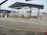 An industrial setting with multiple filling stations under a metal canopy. There are several pipes, valves, and ladders associated with each station. Traffic cones are placed strategically on the ground, and part of the background shows a ship in a dockyard with some cranes visible, indicating a port environment.
