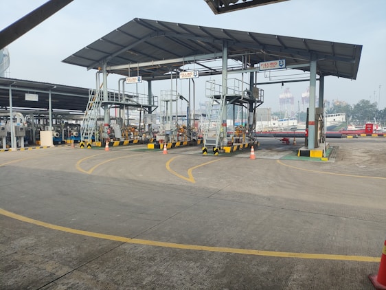 An industrial setting with multiple filling stations under a metal canopy. There are several pipes, valves, and ladders associated with each station. Traffic cones are placed strategically on the ground, and part of the background shows a ship in a dockyard with some cranes visible, indicating a port environment.