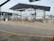 An industrial setting with multiple filling stations under a metal canopy. There are several pipes, valves, and ladders associated with each station. Traffic cones are placed strategically on the ground, and part of the background shows a ship in a dockyard with some cranes visible, indicating a port environment.