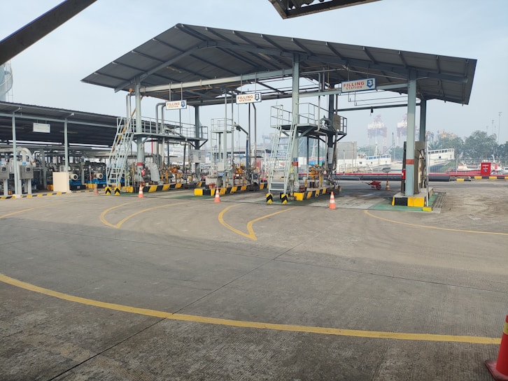An industrial setting with multiple filling stations under a metal canopy. There are several pipes, valves, and ladders associated with each station. Traffic cones are placed strategically on the ground, and part of the background shows a ship in a dockyard with some cranes visible, indicating a port environment.