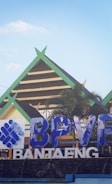 A building with traditional architectural features is seen with a green and brown gabled roof. In the foreground, there is a large blue and white sign displaying the letters 'BPVF' and 'BANTAENG'. A palm tree adds a touch of greenery to the scene, which is set against a clear blue sky.