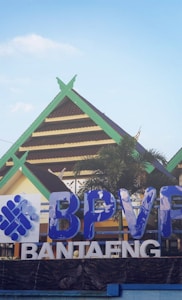 A building with traditional architectural features is seen with a green and brown gabled roof. In the foreground, there is a large blue and white sign displaying the letters 'BPVF' and 'BANTAENG'. A palm tree adds a touch of greenery to the scene, which is set against a clear blue sky.