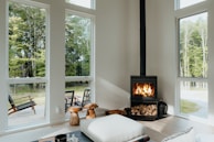 A cozy, modern interior living room with large windows showcasing an outdoor view of lush green trees. A black wood-burning stove, stacked with firewood underneath, emits a warm glow from the flames. Natural light illuminates the room, highlighting minimalistic, comfortable furnishings like a large cushion and wooden stools. Outside, a wooden deck features chairs for relaxation, adding to the serene atmosphere.