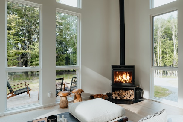 A cozy, modern interior living room with large windows showcasing an outdoor view of lush green trees. A black wood-burning stove, stacked with firewood underneath, emits a warm glow from the flames. Natural light illuminates the room, highlighting minimalistic, comfortable furnishings like a large cushion and wooden stools. Outside, a wooden deck features chairs for relaxation, adding to the serene atmosphere.
