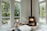 A cozy, modern interior living room with large windows showcasing an outdoor view of lush green trees. A black wood-burning stove, stacked with firewood underneath, emits a warm glow from the flames. Natural light illuminates the room, highlighting minimalistic, comfortable furnishings like a large cushion and wooden stools. Outside, a wooden deck features chairs for relaxation, adding to the serene atmosphere.