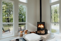 A cozy, modern interior living room with large windows showcasing an outdoor view of lush green trees. A black wood-burning stove, stacked with firewood underneath, emits a warm glow from the flames. Natural light illuminates the room, highlighting minimalistic, comfortable furnishings like a large cushion and wooden stools. Outside, a wooden deck features chairs for relaxation, adding to the serene atmosphere.