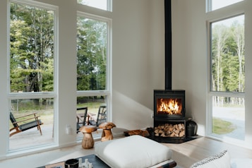 A cozy, modern interior living room with large windows showcasing an outdoor view of lush green trees. A black wood-burning stove, stacked with firewood underneath, emits a warm glow from the flames. Natural light illuminates the room, highlighting minimalistic, comfortable furnishings like a large cushion and wooden stools. Outside, a wooden deck features chairs for relaxation, adding to the serene atmosphere.