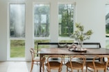 A dining area with large windows that allow ample natural light to flow in, creating a bright and airy space. There is a wooden dining table surrounded by bentwood chairs, topped with a decorative centerpiece featuring a glass vase with green foliage. The view outside the windows shows a lush green landscape.