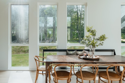 A dining area with large windows that allow ample natural light to flow in, creating a bright and airy space. There is a wooden dining table surrounded by bentwood chairs, topped with a decorative centerpiece featuring a glass vase with green foliage. The view outside the windows shows a lush green landscape.
