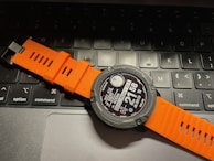 A digital smartwatch with an orange strap is resting on a laptop keyboard. The watch face displays various metrics and information, and the keyboard keys are visible beneath it.
